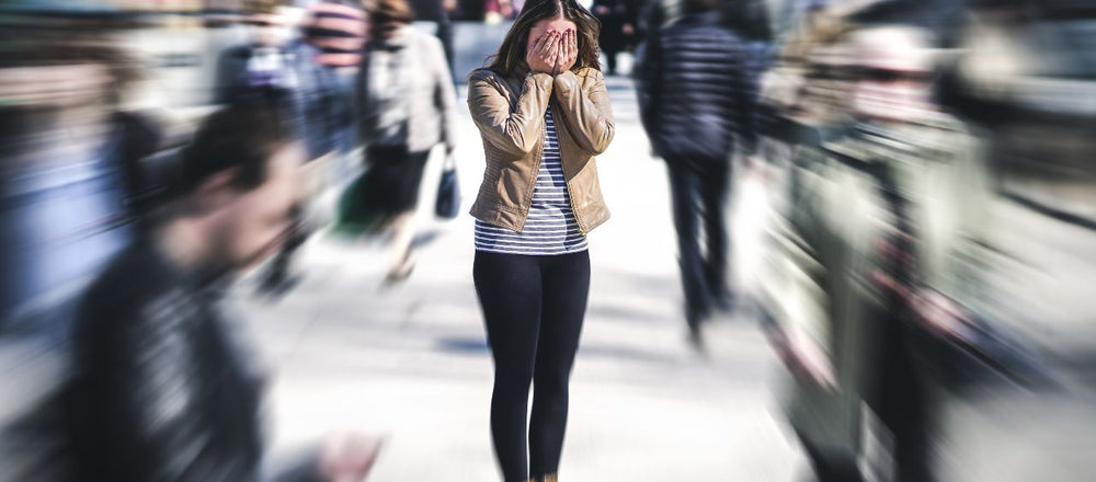 Woman experiencing anxiety as she stands with hands on her face in the middle of a crowded street with blurred out pedestrians.