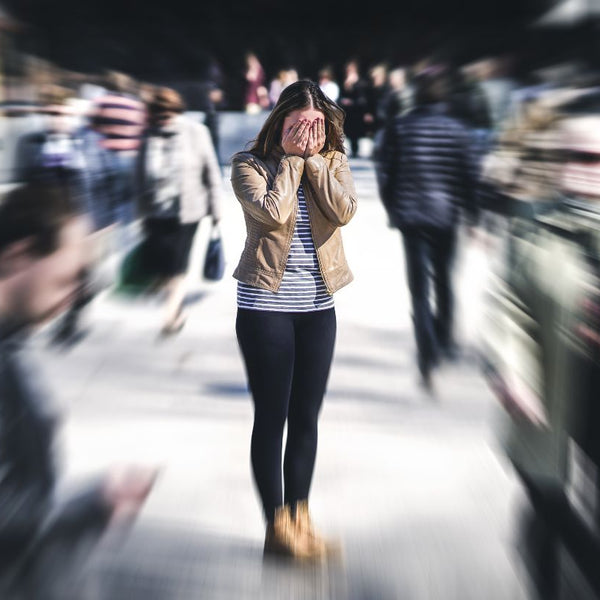 Woman experiencing anxiety as she stands with hands on her face in the middle of a crowded street with blurred out pedestrians.